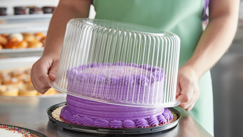 Baker placing a clear plastic cake dome over a purple buttercream-frosted cake to store it properly