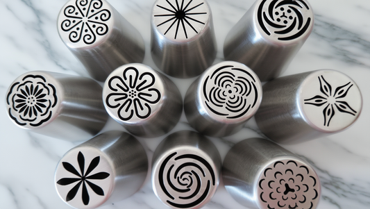 Collection of stainless steel Russian piping tips arranged on marble, each showing a different flower and swirl pattern on its opening